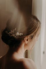 La femme porte une robe bustier blanche,elle est de dos avec un chignon bas flou et porte un peigne de fleurs mariage mariée en porcelaine