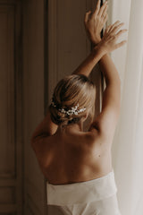 La femme porte une robe bustier blanche,elle est de dos avec un chignon bas flou et porte un peigne de fleurs mariage mariée en porcelaine