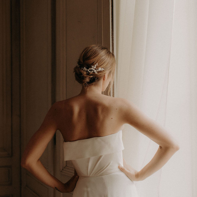 La femme porte une robe bustier blanche,elle est de dos avec un chignon bas flou et porte un peigne de fleurs mariage mariée en porcelaine