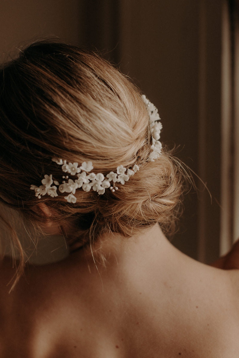 La femme porte une robe bustier blanche,elle est de dos avec un chignon bas flou et porte un peigne de fleurs mariage mariée en porcelaine